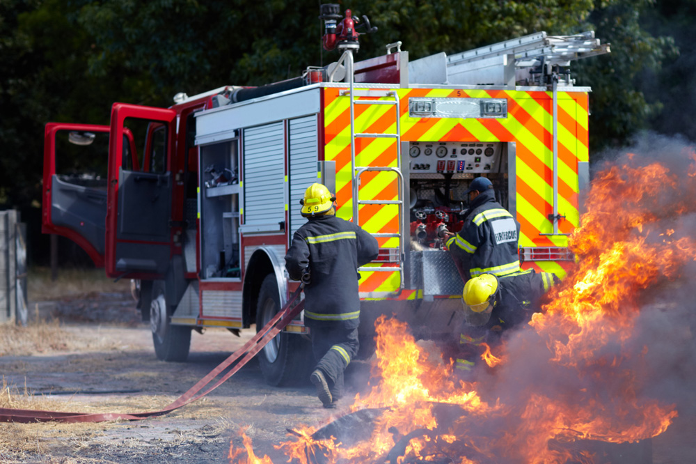 Brandbil i bakgrunden med flera brandmän runtomkring medan det brinner i förgrunden.